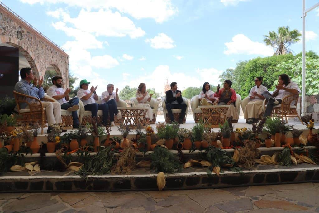 Inicia el Festival Endémico 2025 que muestra lo mejor de la gastronomía de Guanajuato Personas participando en panel de discusión con decoración de flores y plantas, bajo cielo despejado en evento cultural.