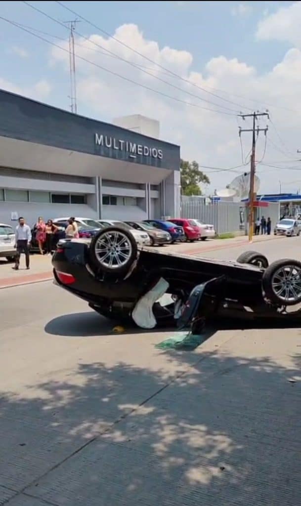 Accidente de coche volcado frente a edificio de multimédios, impacto automovilístico grave en la vía pública.