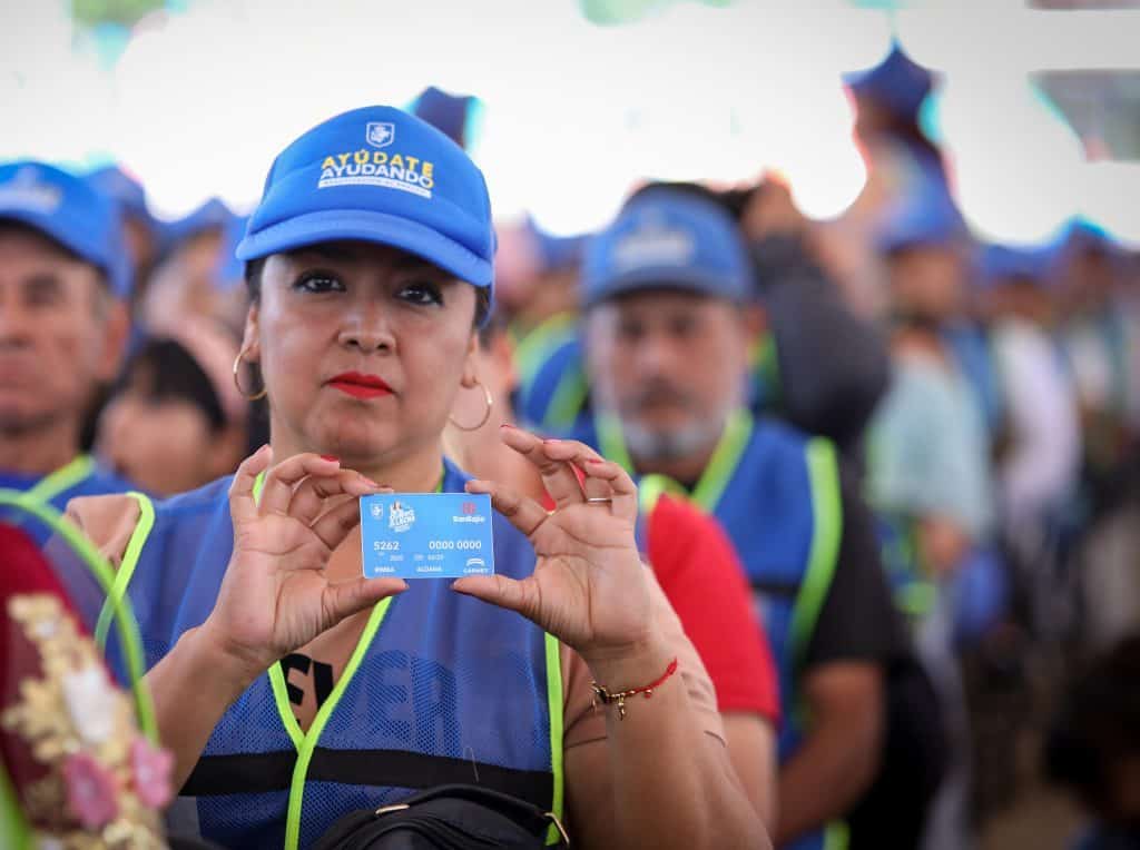 Alto: Mujer con gorra azul y vestimenta de ayuda sostiene tarjeta de donación en evento solidario.