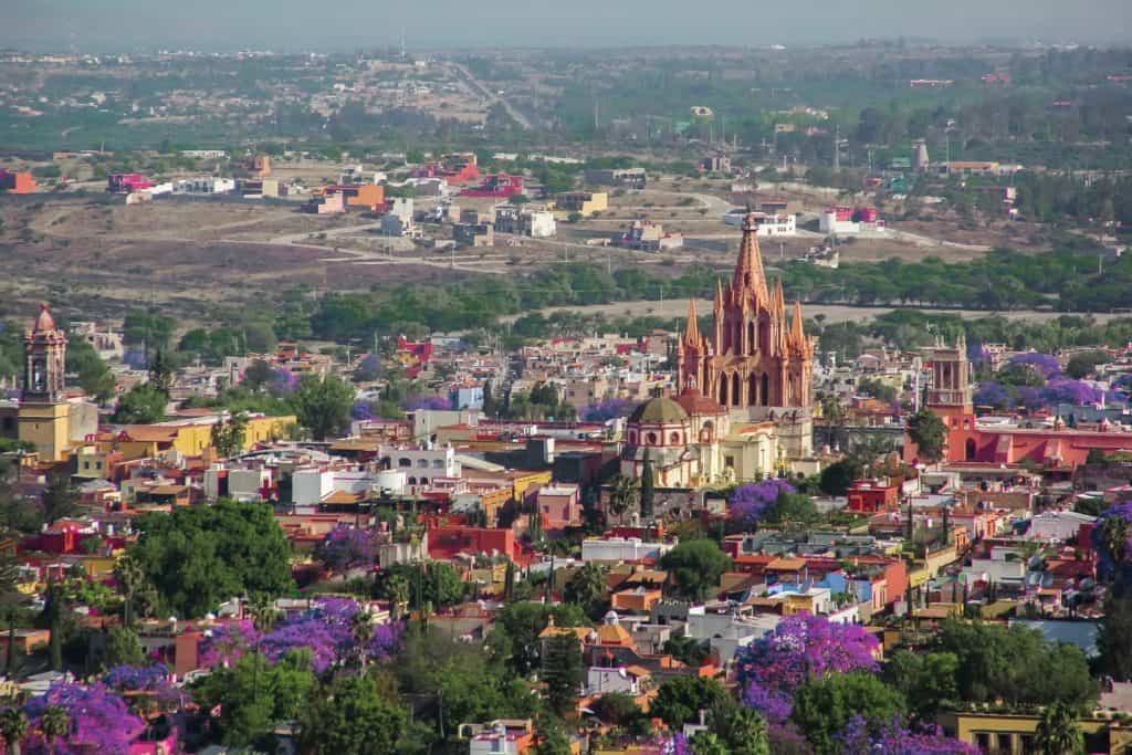 Catedral en la ciudad de San Miguel de Allende, rodeada de casas coloridas y árboles con jacarandas en flor.