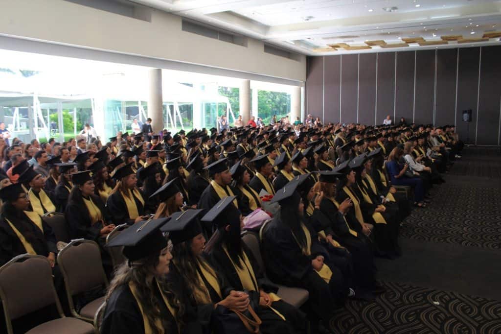 Felicidades a todos los graduados en ceremonia académica en un salón con gran aforo y ventanales con vista al exterior.