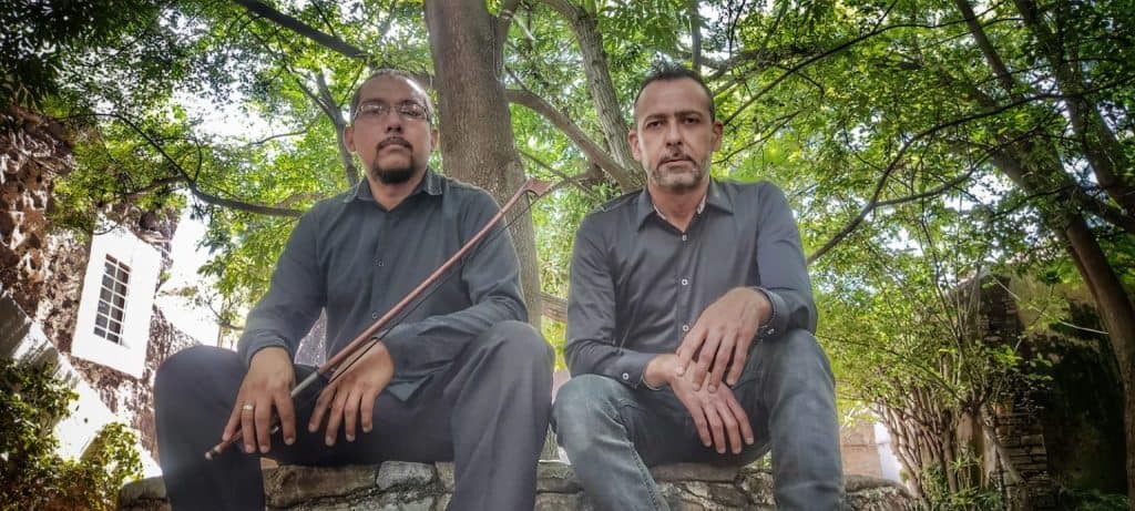 Hombres con camisas negras en un entorno natural con árboles y paredes de piedra.