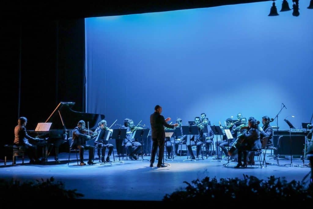 Refuerzo presencial de orquesta en escenario con iluminación azul, dirección de un conductor, música en vivo, evento cultural.