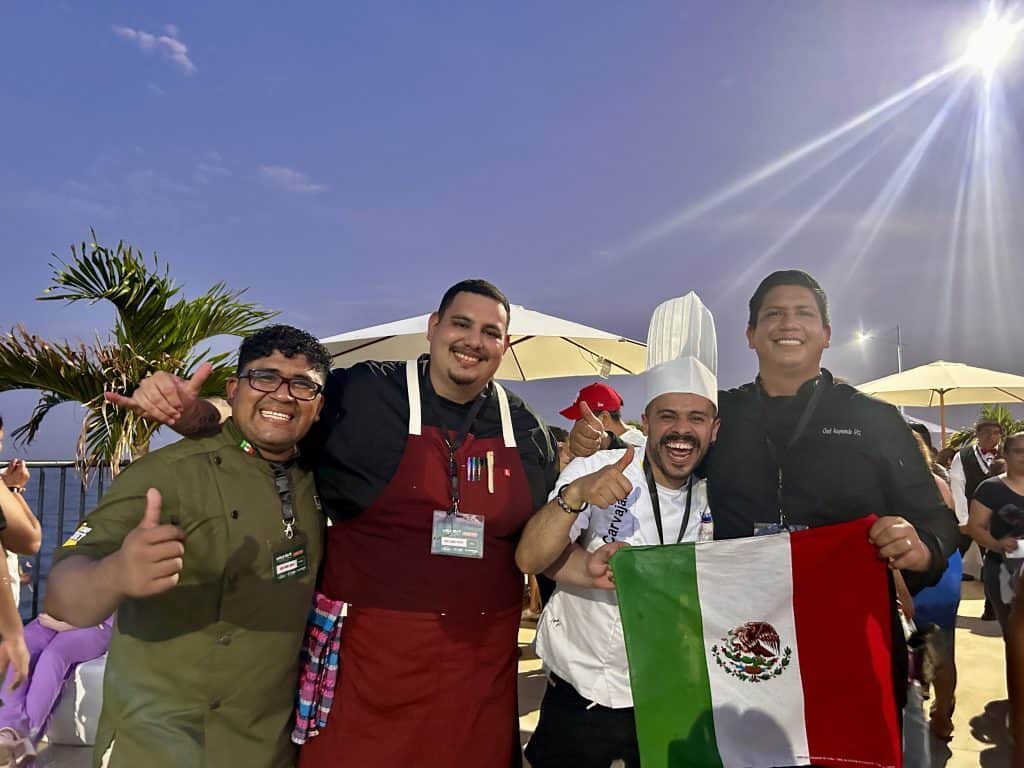 Personas festejando con bandera de México en evento gastronómico al aire libre bajo el sol brillante en la tarde.