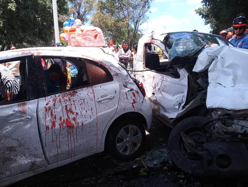 Accidente de coche grave con dos vehículos severamente golpeados, attended by bystanders, en un incidente vial en una calle con árboles.