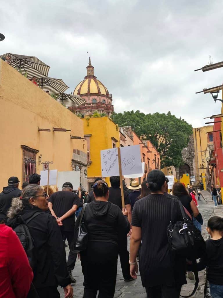 Personas en manifestación en calle con edificios históricos y cúpula en fondo, protestando por justicia y derechos.