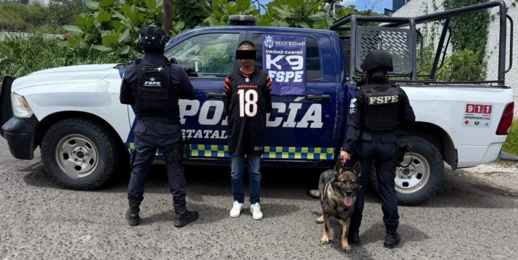 Hombre detenido con oficiales y perro policía frente a camión policial en la calle.