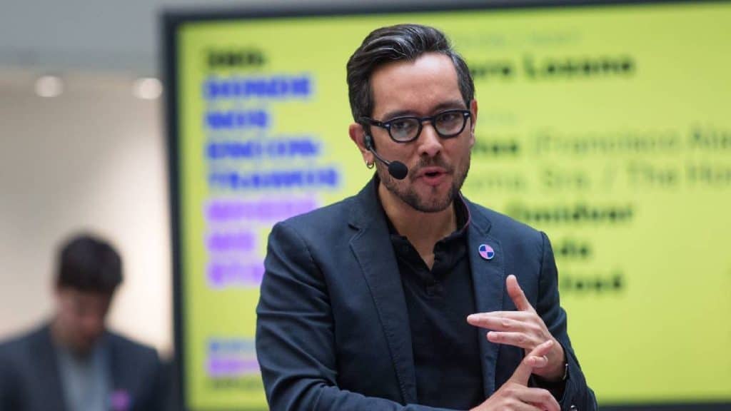 Hombre en conferencia con micrófono, usando blazer y gafas, en entorno profesional y con pantalla de fondo.