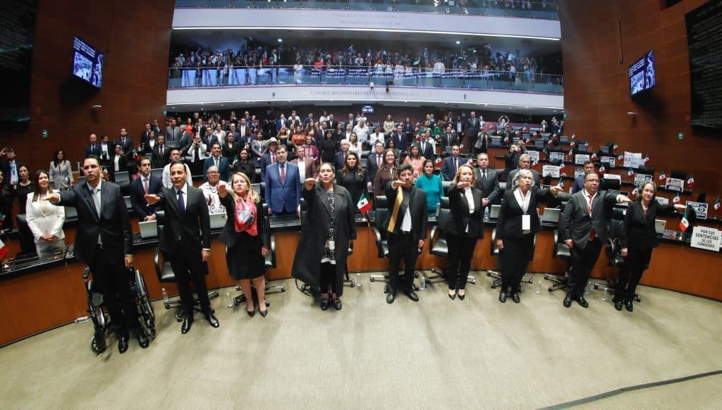 Hombre y mujeres en sesión del Congreso Mexicano, levantando la mano en juramento.