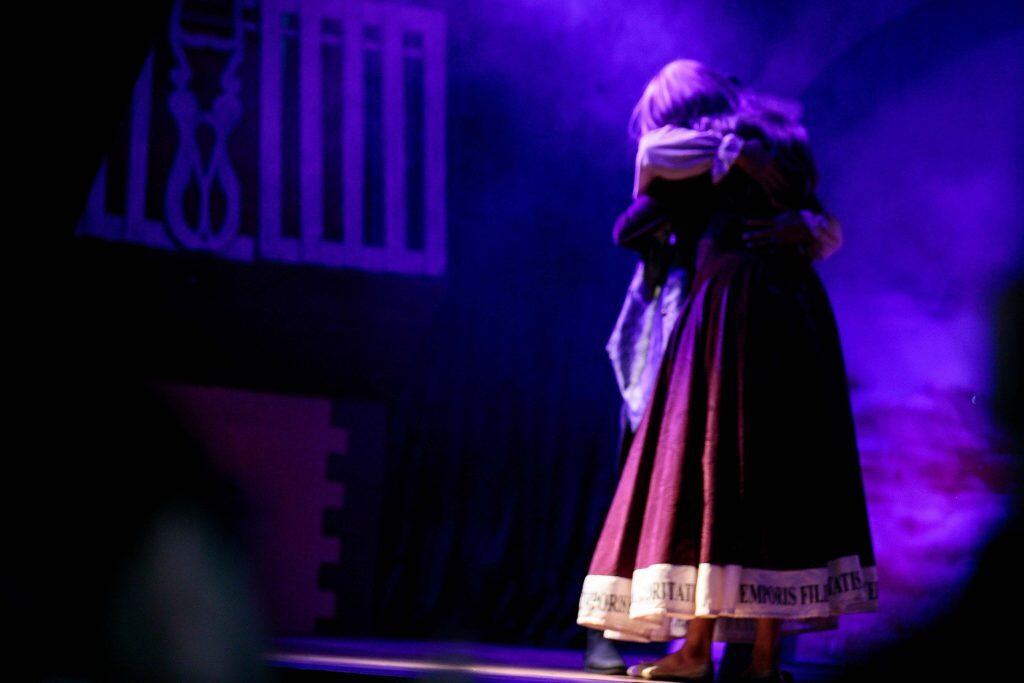 Vestuario tradicional en obra teatral con iluminación violeta, personajes abrazados en escena cultural.