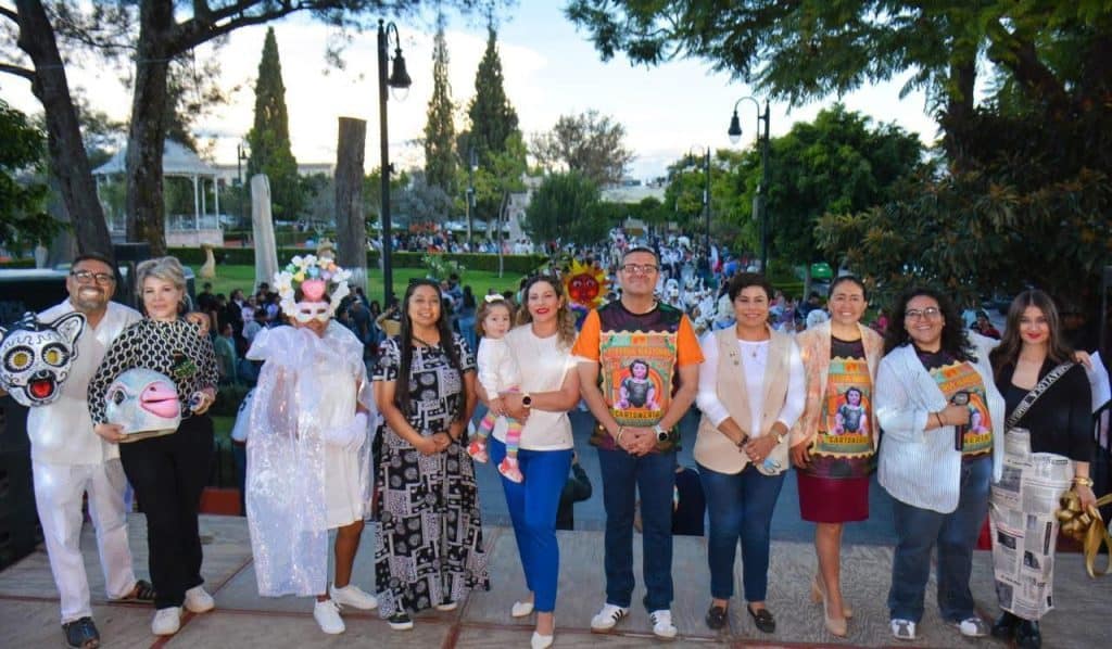Diversos grupos en evento cultural en parque, con disfraces y arte popular mexicano.