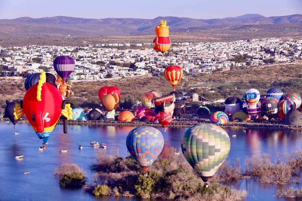 Coloresidos globos de hot air en festival en la ciudad con montañas de fondo y agua en primer plano.