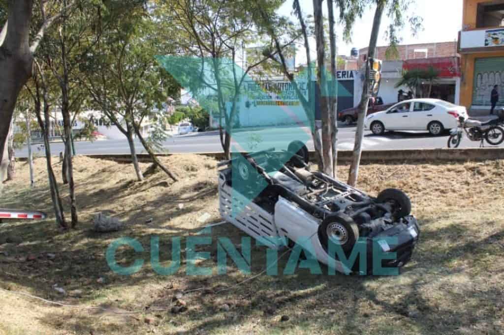 Accidente de coche volcado en la calle con árboles y tráfico en fondo.
