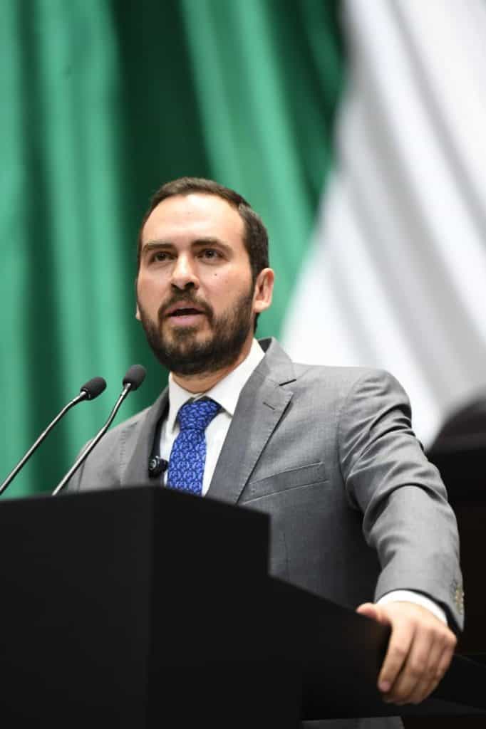 Hombre en acto de hablar en un escenario con fondo de banderas, vestimenta formal.