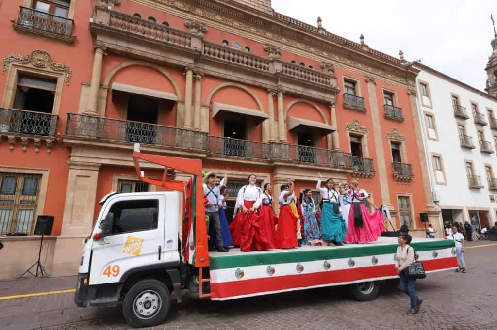 Personas en vestimenta tradicional mexicana en desfile con camioneta decorada con banderas de México.