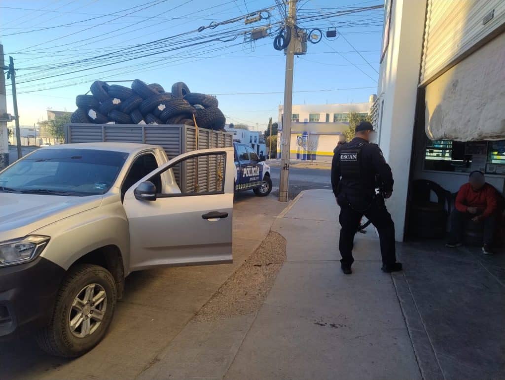 Robo de llantas de neumáticos en vehículo en la calle, presencia de policía y testigo sentado en banco.