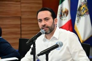 Alto de 100 caracteres: político mexicano en conferencia con banderas de México y estados en el fondo.