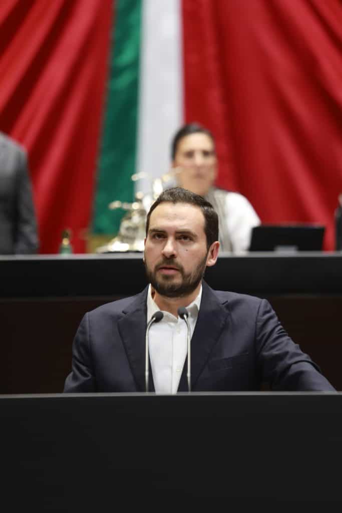 Diputado Alan Márquez pide reforzar la seguridad en carreteras federales Representante en conferencia con banderas mexicanas, expresión seria en evento político o social.