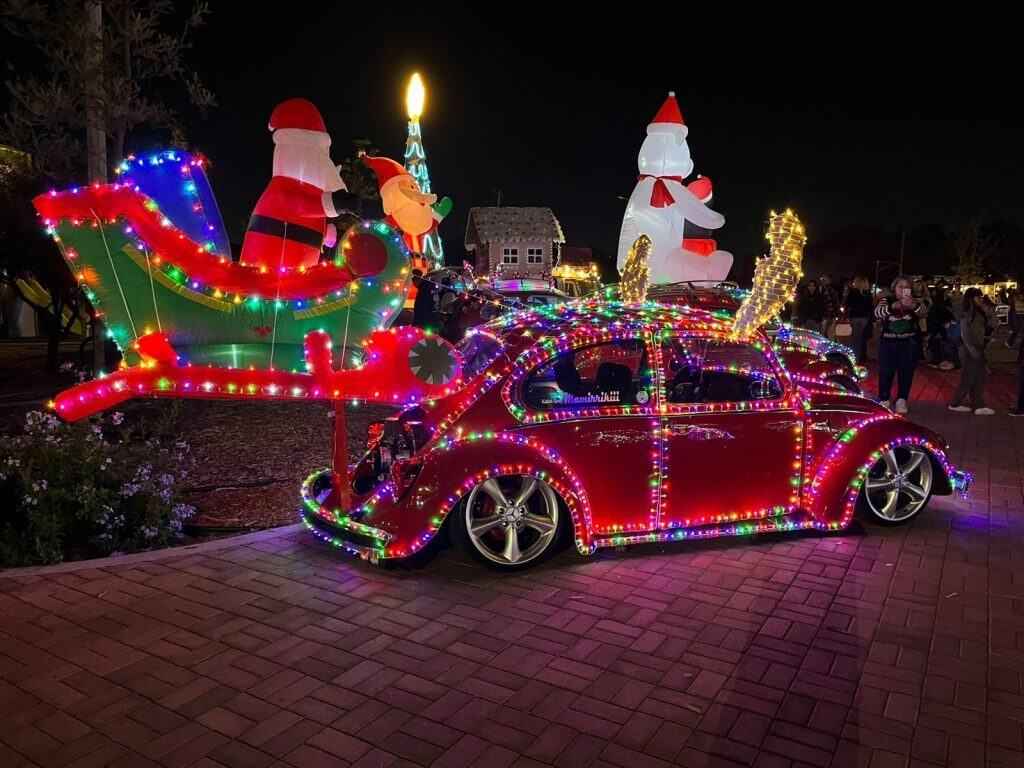 Luces navideñas en un coche decorado con motivos festivos, Santa Claus y reno en un evento navideño al aire libre.