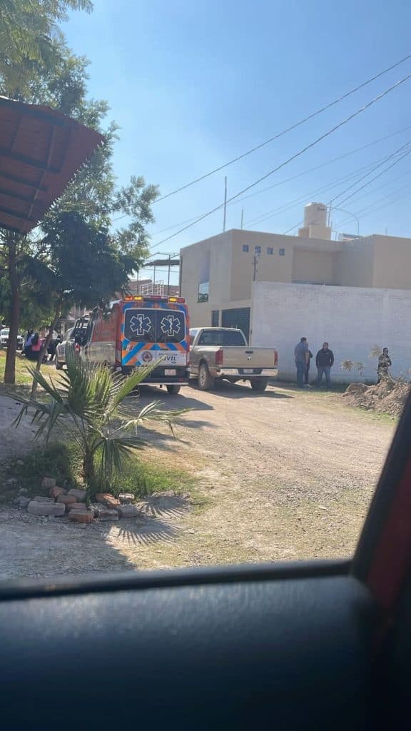 Ambulancia en la calle con paramédicos y personas en la escena, en una zona urbana con vivienda y árboles.