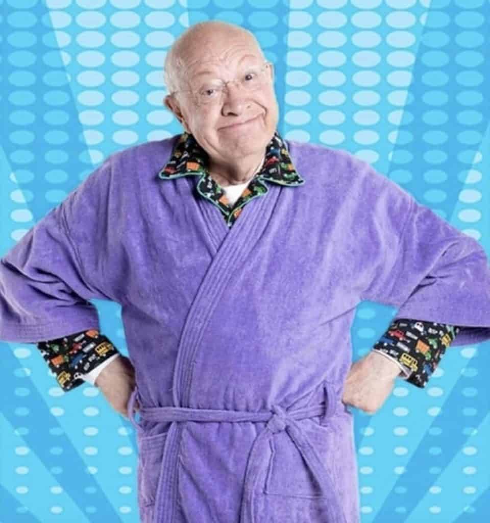 Divertido anciano con bata morada y camisa de personajes de dibujos animados, sonrisa confiada y actitud amigable.