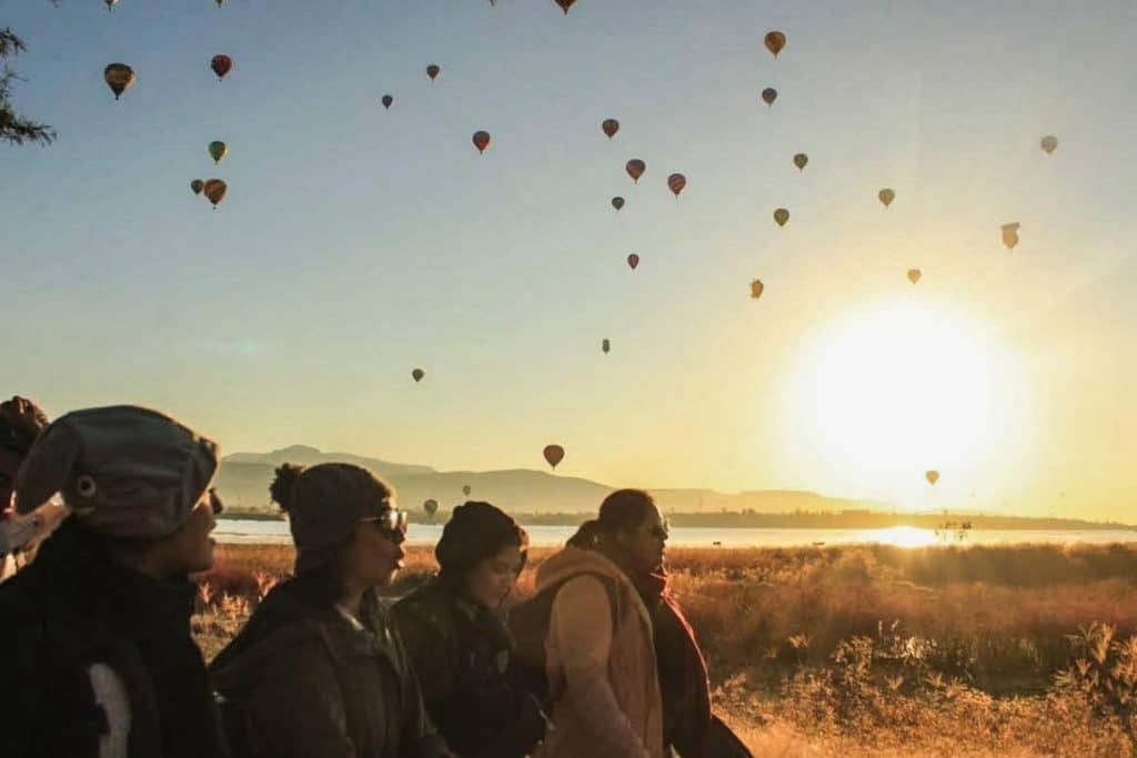 Personas apreciando globos aerostáticos al atardecer en un paisaje natural/turístico.