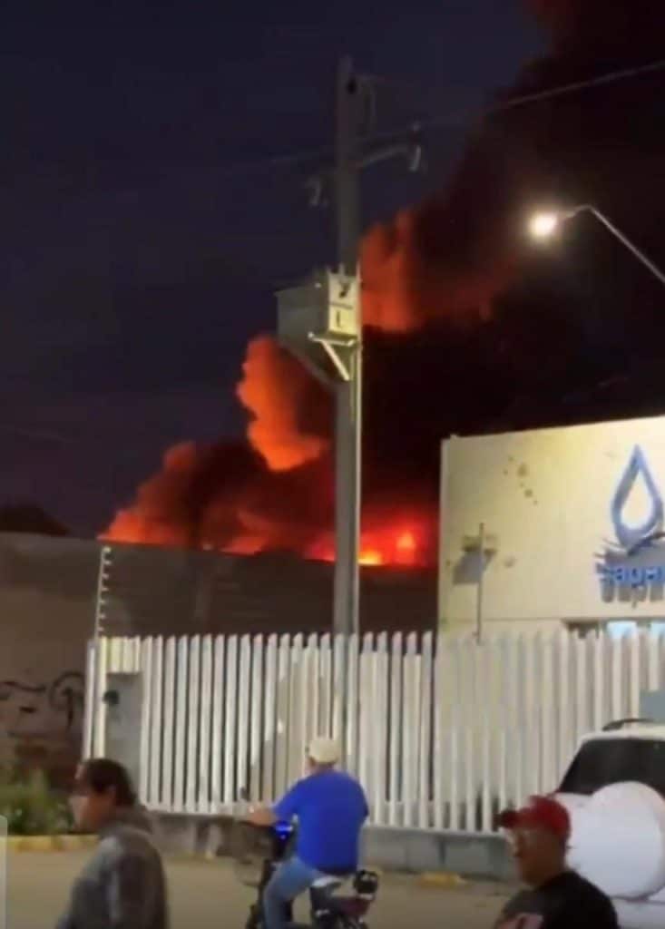 Atienden bomberos incendio en recicladora de la colonia Santa María del Granjeno Incendio en una zona urbana con humo y fuego visibles en la noche, personas observan la emergencia desde la calle.
