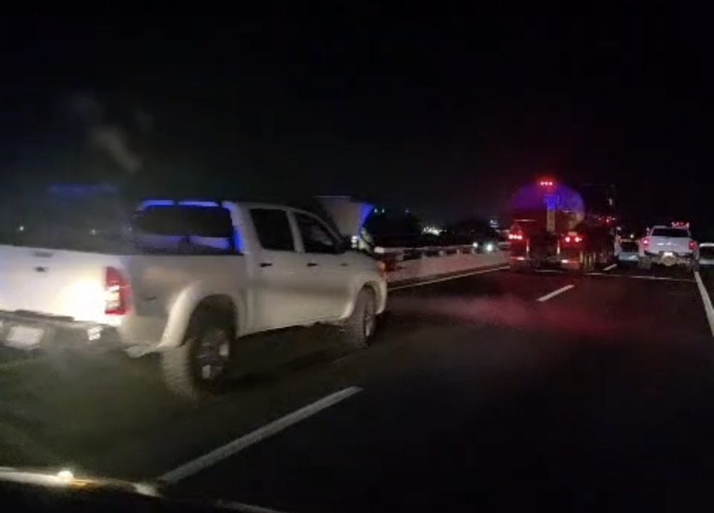 Camión de servicios en carretera durante la noche, luces rojas y vehículos en fila, en una noche oscura.