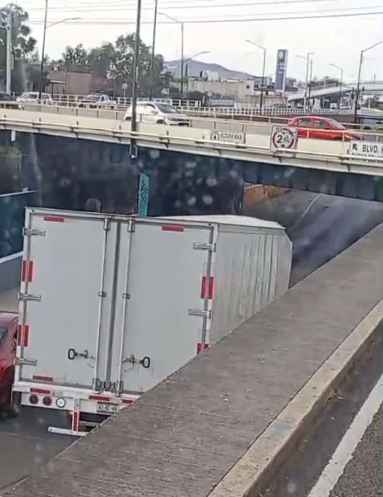 Camión de carga en semáforo en ciudad con tráfico y puente elevado en fondo.