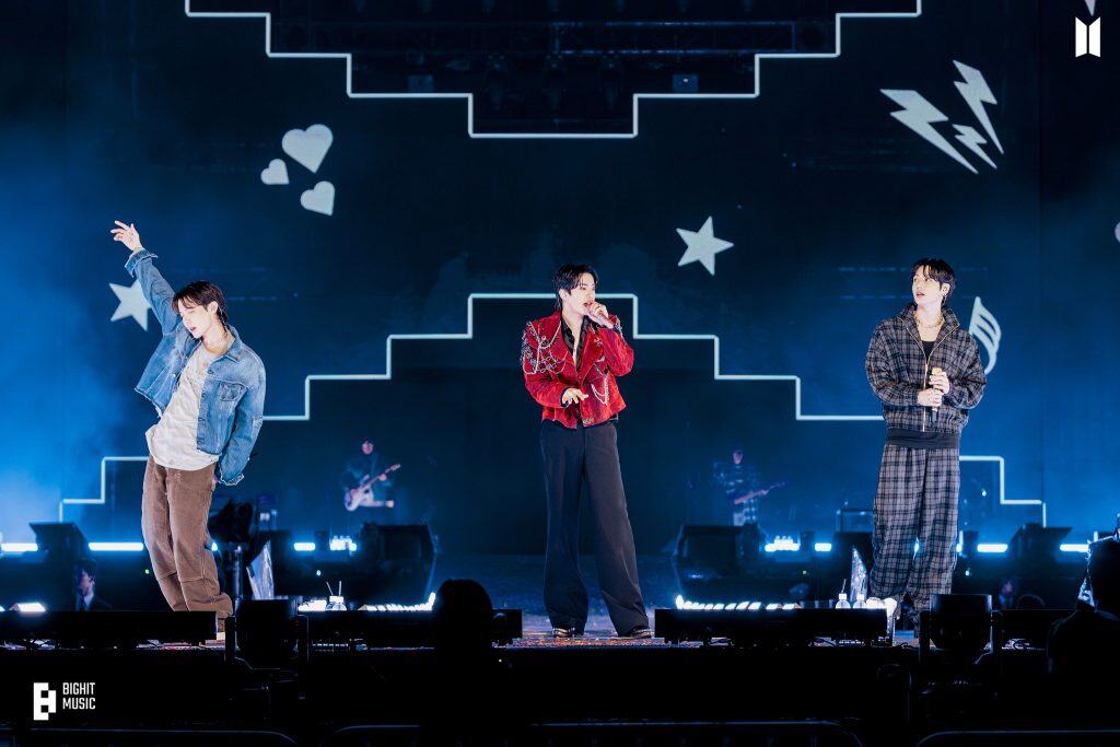 Jóvenes cantantes en concierto con escenario iluminado y diseño de estrellas y corazones en el fondo.